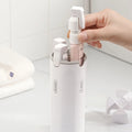 Hand holding a white spray bottle with a pink pump over a white container with multiple spray bottles.